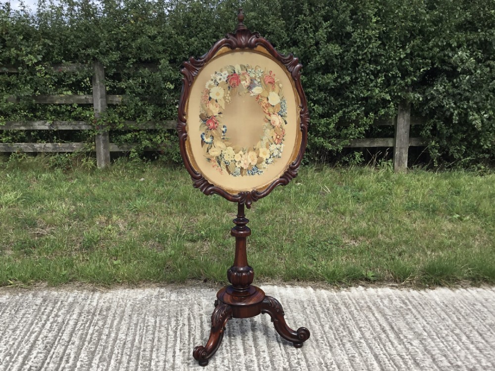 An antique Victorian rosewood pole screen featuring a carved oval frame with a floral wreath needlepoint panel, supported on a turned column with a tripod cabriole base.
