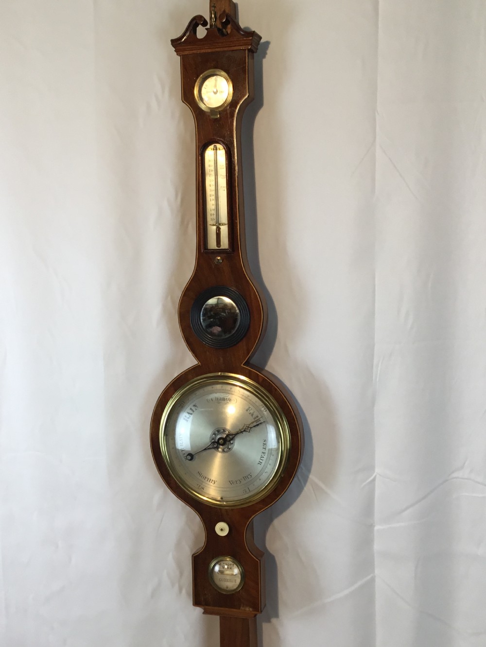 An antique mahogany banjo-shaped wheel barometer featuring a broken arch pediment, hygrometer, thermometer, convex mirror, and silvered dial.