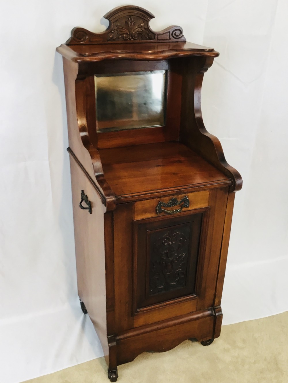 A late Victorian carved wooden coal purdonium featuring an ornate upper crest, a small mirrored back shelf, and a drop-front cabinet door with a brass handle.