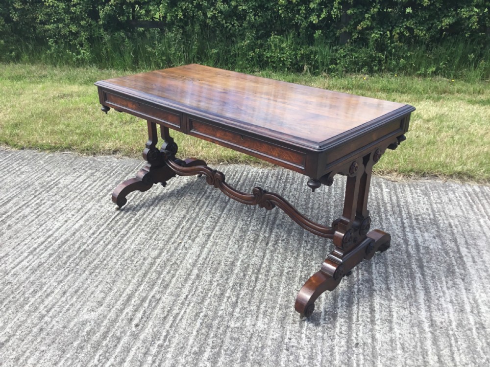 A William IV rosewood library table featuring a rectangular top, frieze drawers, turned trestle end supports, a carved stretcher, and scroll feet on castors.