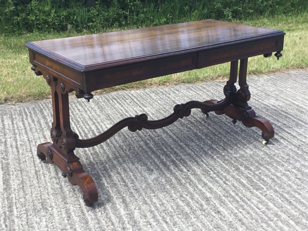 A William IV rosewood library table featuring a rectangular top, frieze drawers, turned trestle end supports, a carved stretcher, and scroll feet on castors. View III