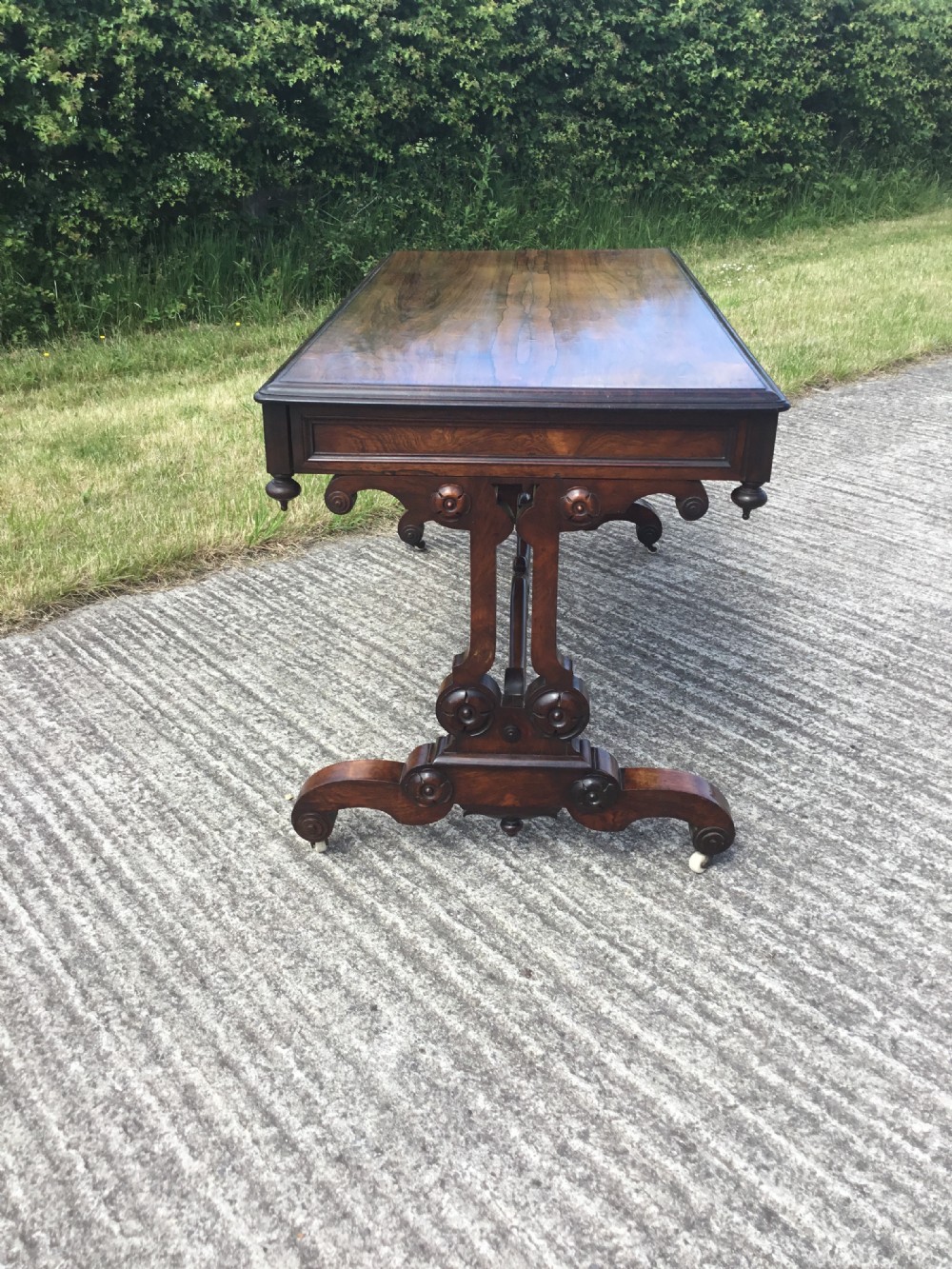 A William IV rosewood library table featuring a rectangular top, frieze drawers, turned trestle end supports, a carved stretcher, and scroll feet on castors. View V