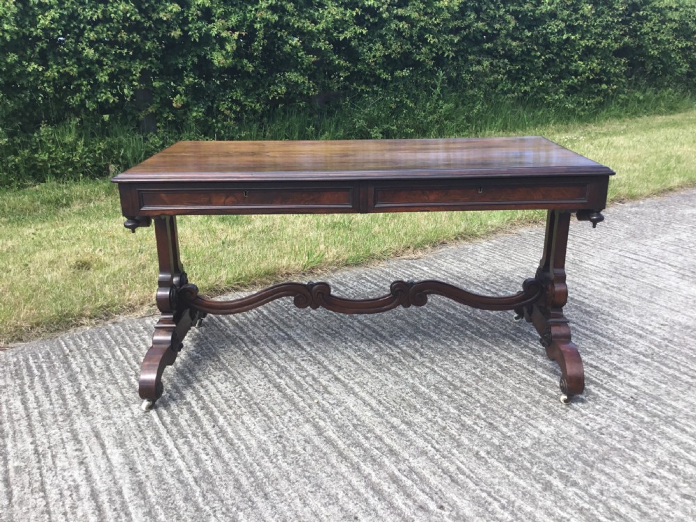 A William IV rosewood library table featuring a rectangular top, frieze drawers, turned trestle end supports, a carved stretcher, and scroll feet on castors. View VI