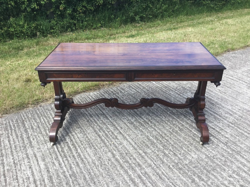 A William IV rosewood library table featuring a rectangular top, frieze drawers, turned trestle end supports, a carved stretcher, and scroll feet on castors. View VII