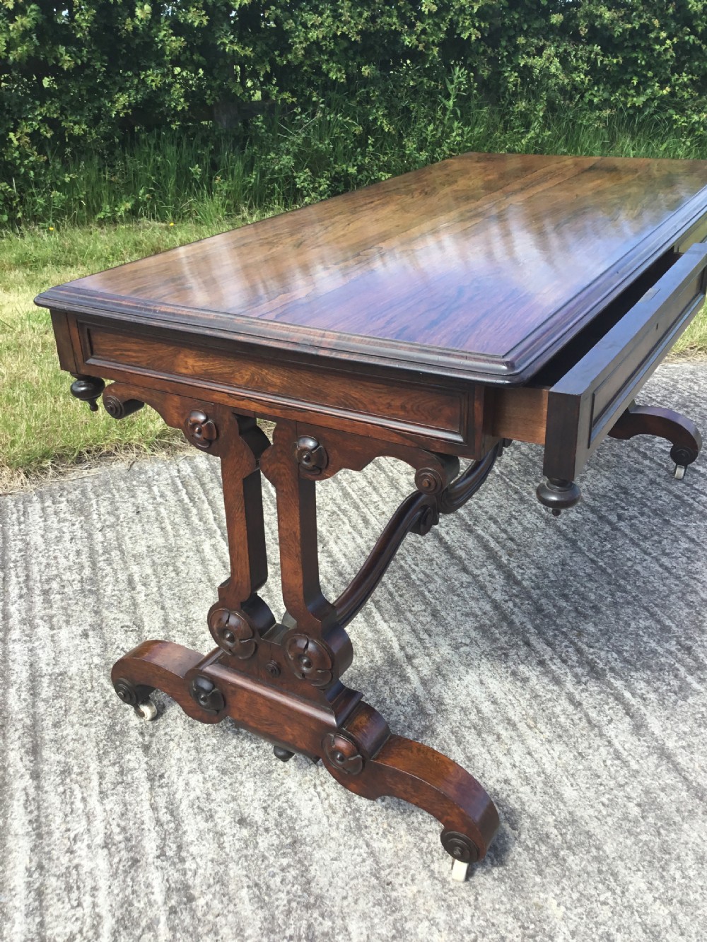 A William IV rosewood library table featuring a rectangular top, frieze drawers, turned trestle end supports, a carved stretcher, and scroll feet on castors. View X