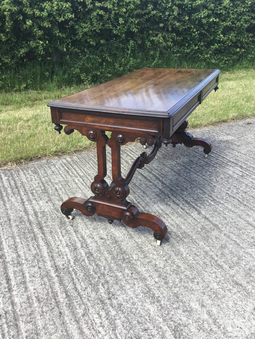 A William IV rosewood library table featuring a rectangular top, frieze drawers, turned trestle end supports, a carved stretcher, and scroll feet on castors. View XII