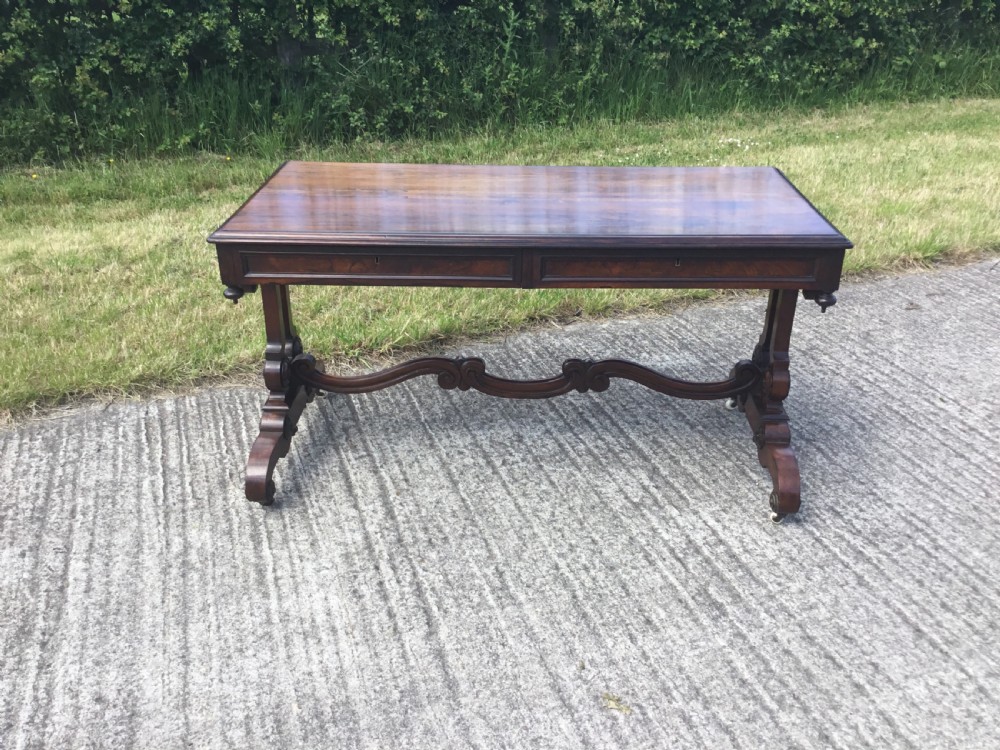 A William IV rosewood library table featuring a rectangular top, frieze drawers, turned trestle end supports, a carved stretcher, and scroll feet on castors. View XIII