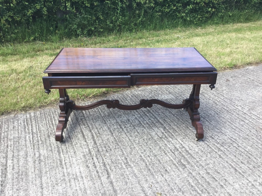 A William IV rosewood library table featuring a rectangular top, frieze drawers, turned trestle end supports, a carved stretcher, and scroll feet on castors. View XIV