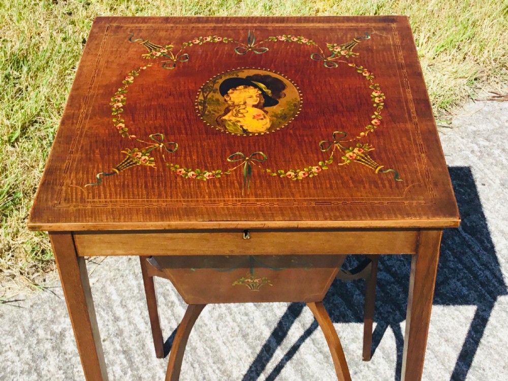 An antique square wooden work table featuring a painted portrait medallion surrounded by floral garlands on the lid, supported by square tapered legs with arched stretchers. View X