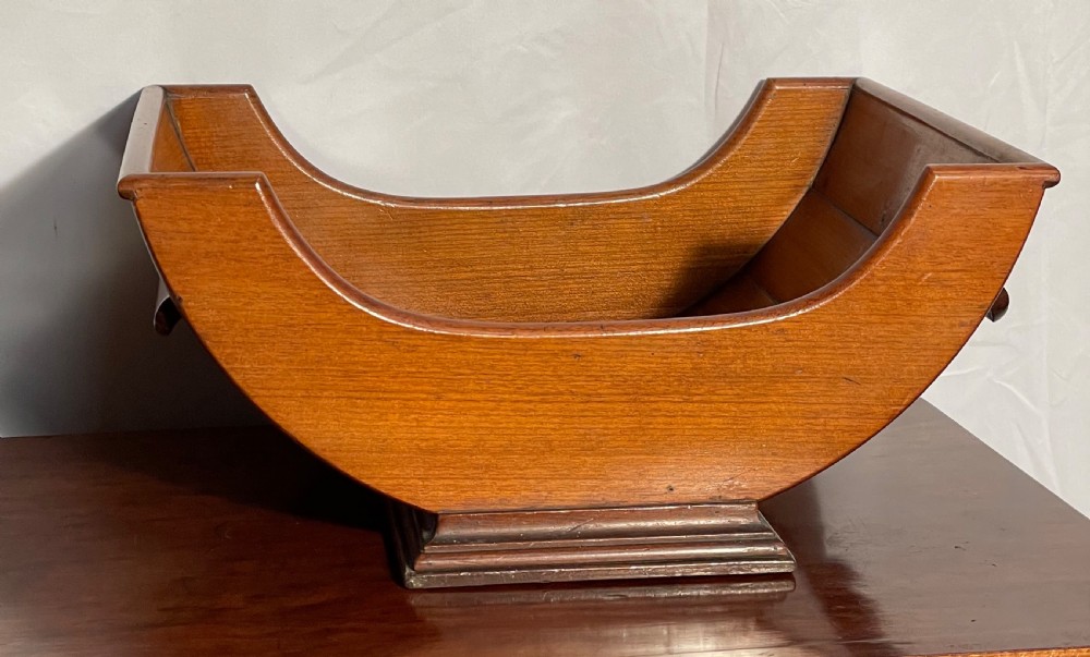 A mahogany wooden cheese cradle featuring a curved slatted trough mounted on a moulded rectangular plinth base. View II