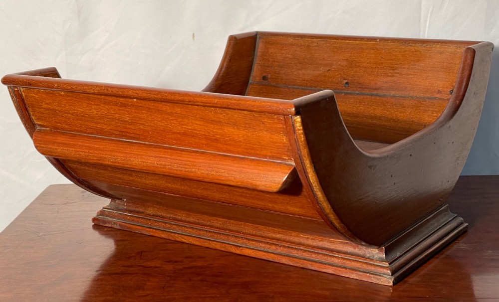 A mahogany wooden cheese cradle featuring a curved slatted trough mounted on a moulded rectangular plinth base. View VI
