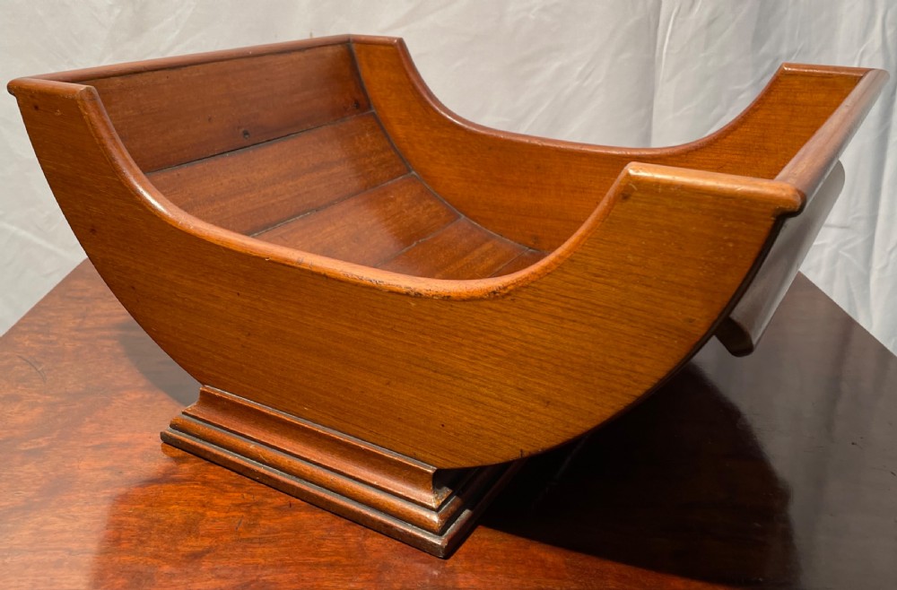 A mahogany wooden cheese cradle featuring a curved slatted trough mounted on a moulded rectangular plinth base. View VII