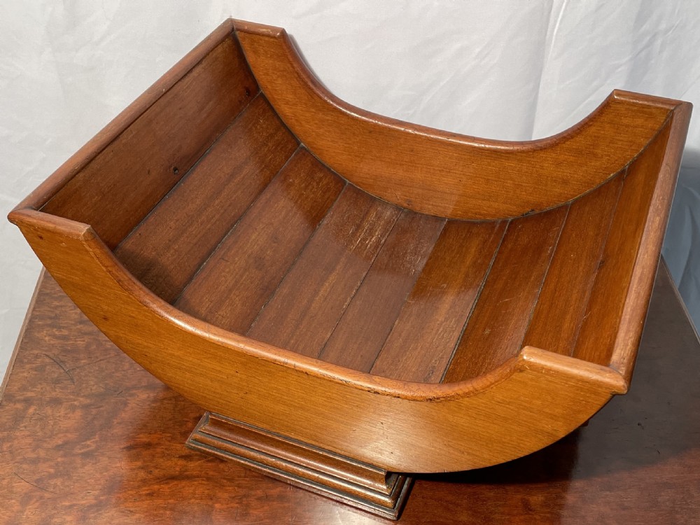 A mahogany wooden cheese cradle featuring a curved slatted trough mounted on a moulded rectangular plinth base. View VIII