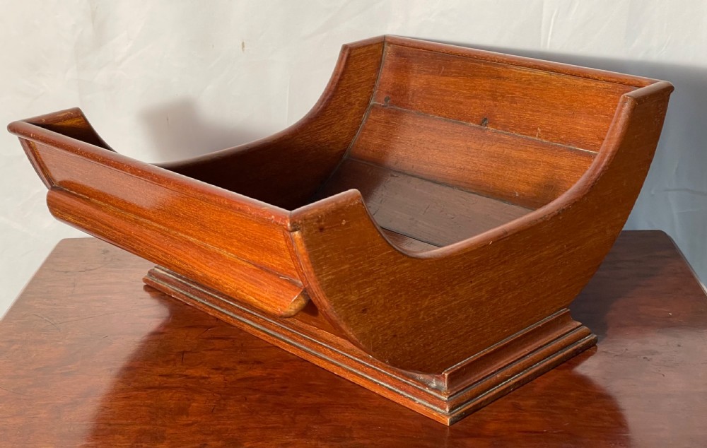 A mahogany wooden cheese cradle featuring a curved slatted trough mounted on a moulded rectangular plinth base. View IX