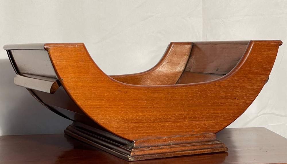 A mahogany wooden cheese cradle featuring a curved slatted trough mounted on a moulded rectangular plinth base. View XI
