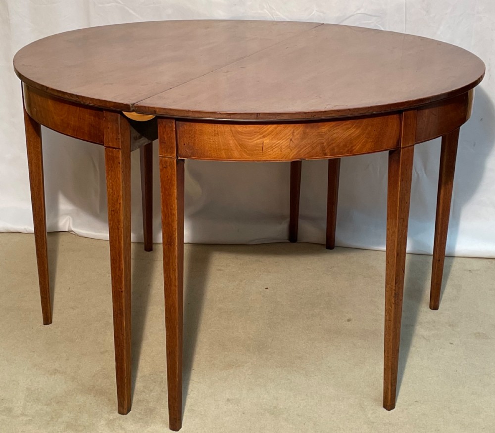 A Georgian mahogany extension dining table featuring a round top assembled from two demi-lune end sections and a central leaf, supported by slender square tapered legs. View X