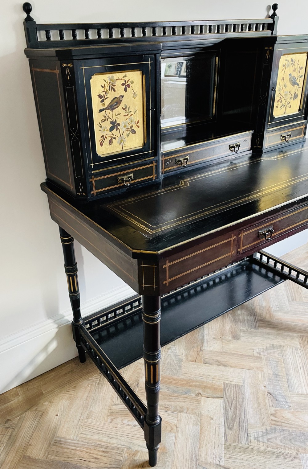 An ebonised Aesthetic Movement bonheur du jour featuring gold line detailing, central mirror, two hand-painted bird panels on small upper cabinets, and turned legs with a spindle lower gallery. View X