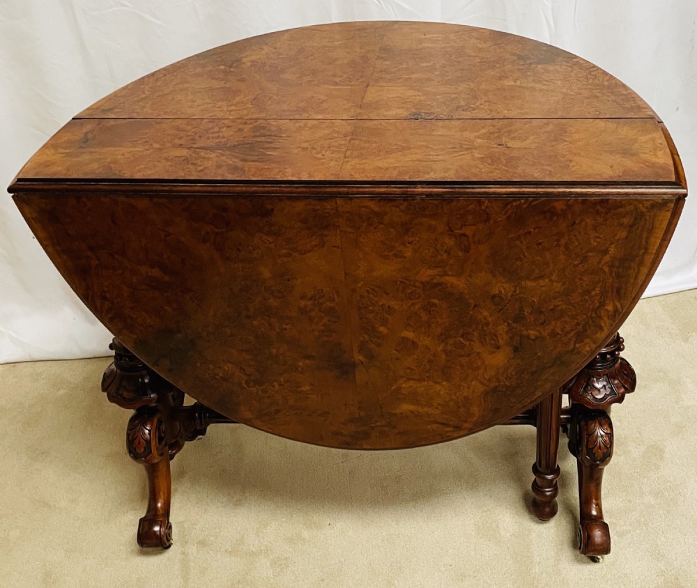 A Victorian burr walnut Sutherland table shown with folded drop leaves, turned end supports, carved cabriole feet, and white ceramic castors. View II