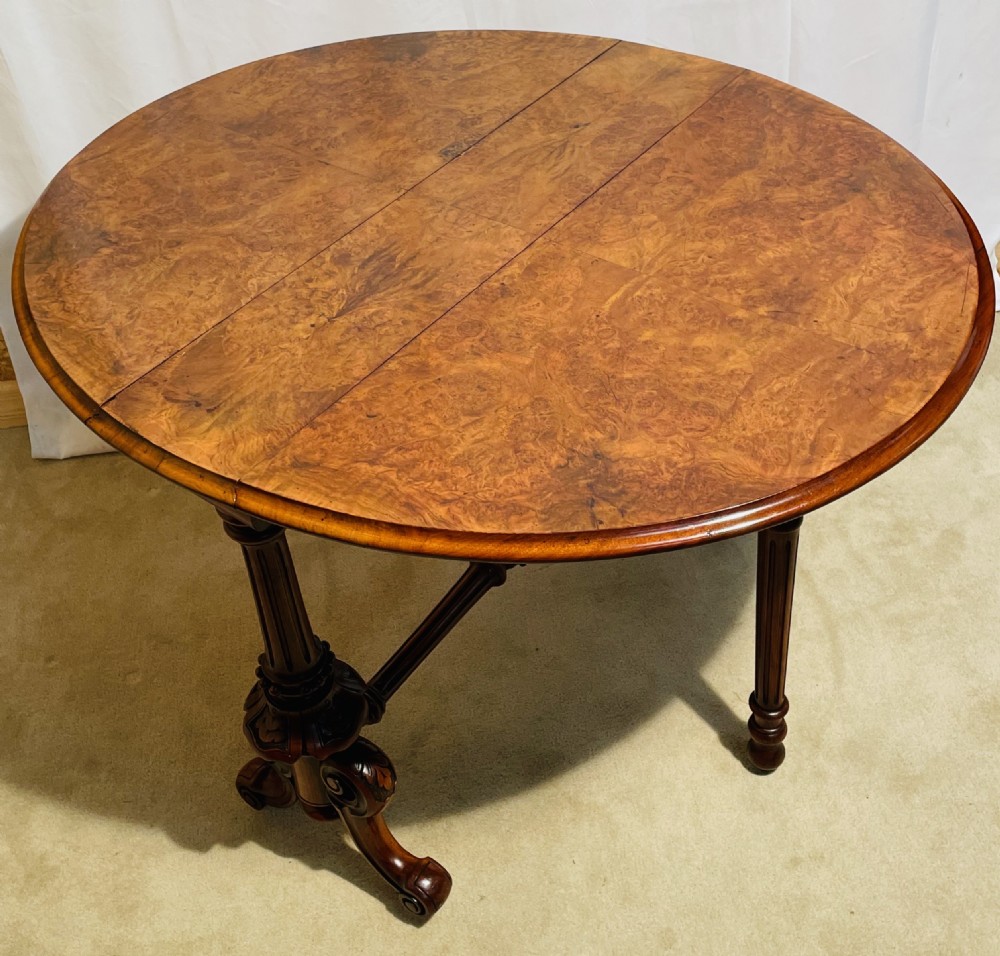 A Victorian burr walnut Sutherland table shown with folded drop leaves, turned end supports, carved cabriole feet, and white ceramic castors. View VI