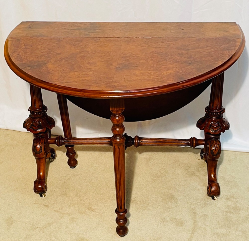 A Victorian burr walnut Sutherland table shown with folded drop leaves, turned end supports, carved cabriole feet, and white ceramic castors. View X