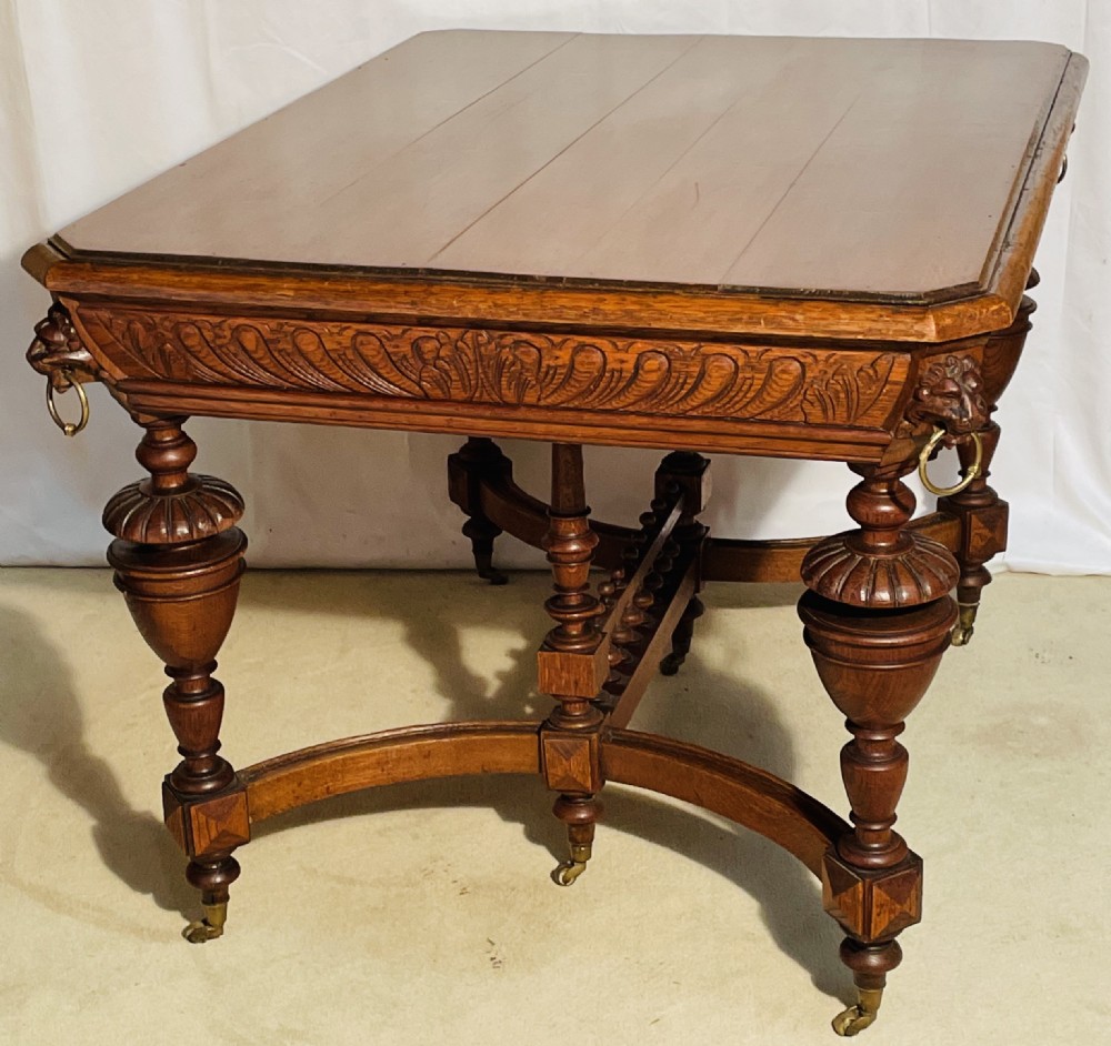 A Victorian Gothic honey oak centre table featuring carved aprons, corner lion mask motifs with brass rings, turned legs, and an arched cross-stretcher base on castors.