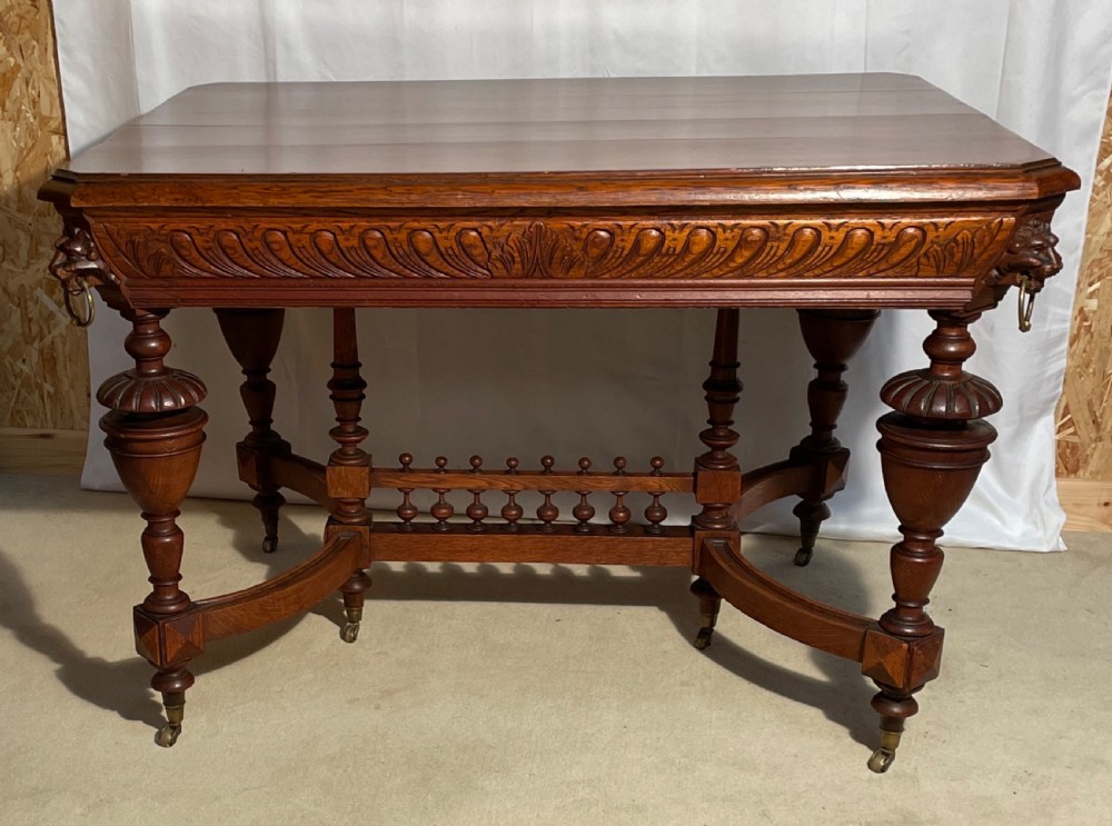 A Victorian Gothic honey oak centre table featuring carved aprons, corner lion mask motifs with brass rings, turned legs, and an arched cross-stretcher base on castors. View III