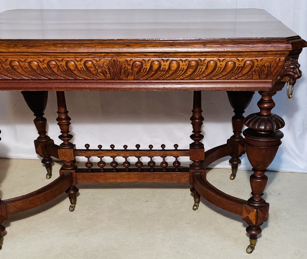 A Victorian Gothic honey oak centre table featuring carved aprons, corner lion mask motifs with brass rings, turned legs, and an arched cross-stretcher base on castors. View IV