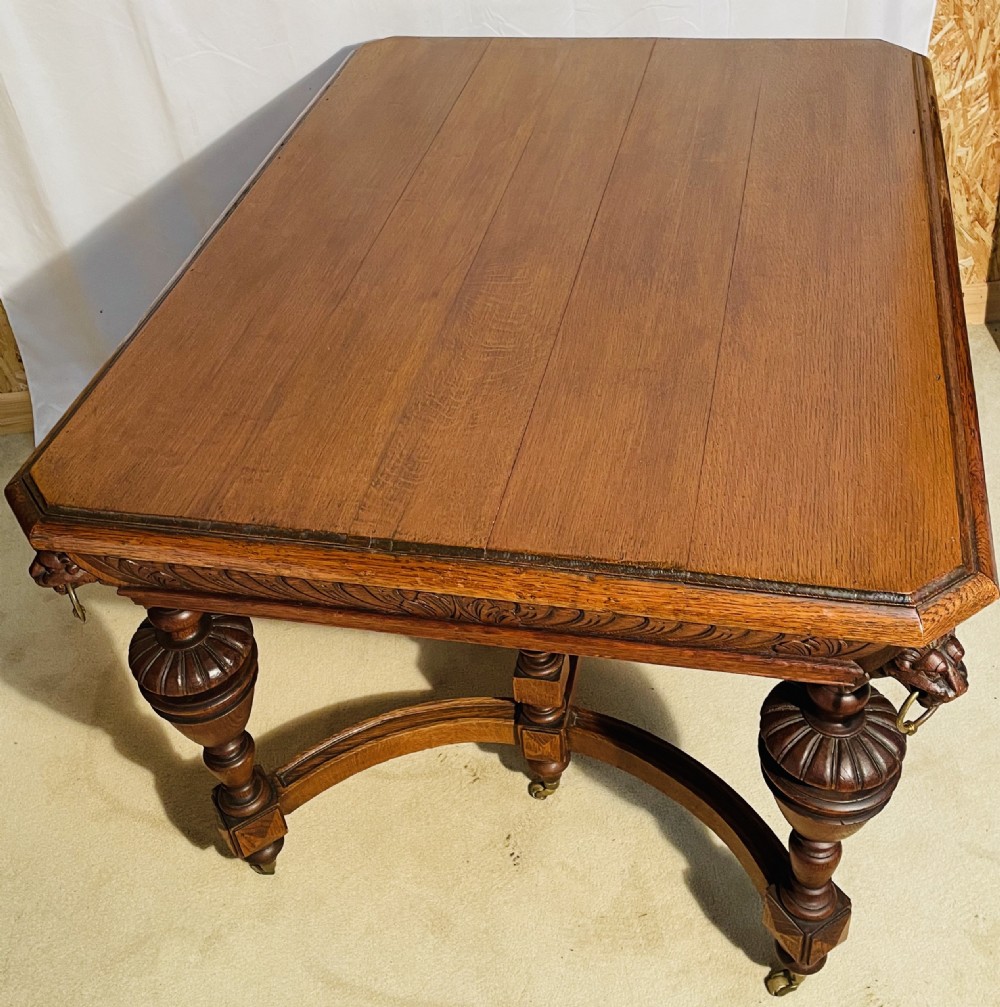 A Victorian Gothic honey oak centre table featuring carved aprons, corner lion mask motifs with brass rings, turned legs, and an arched cross-stretcher base on castors. View VI