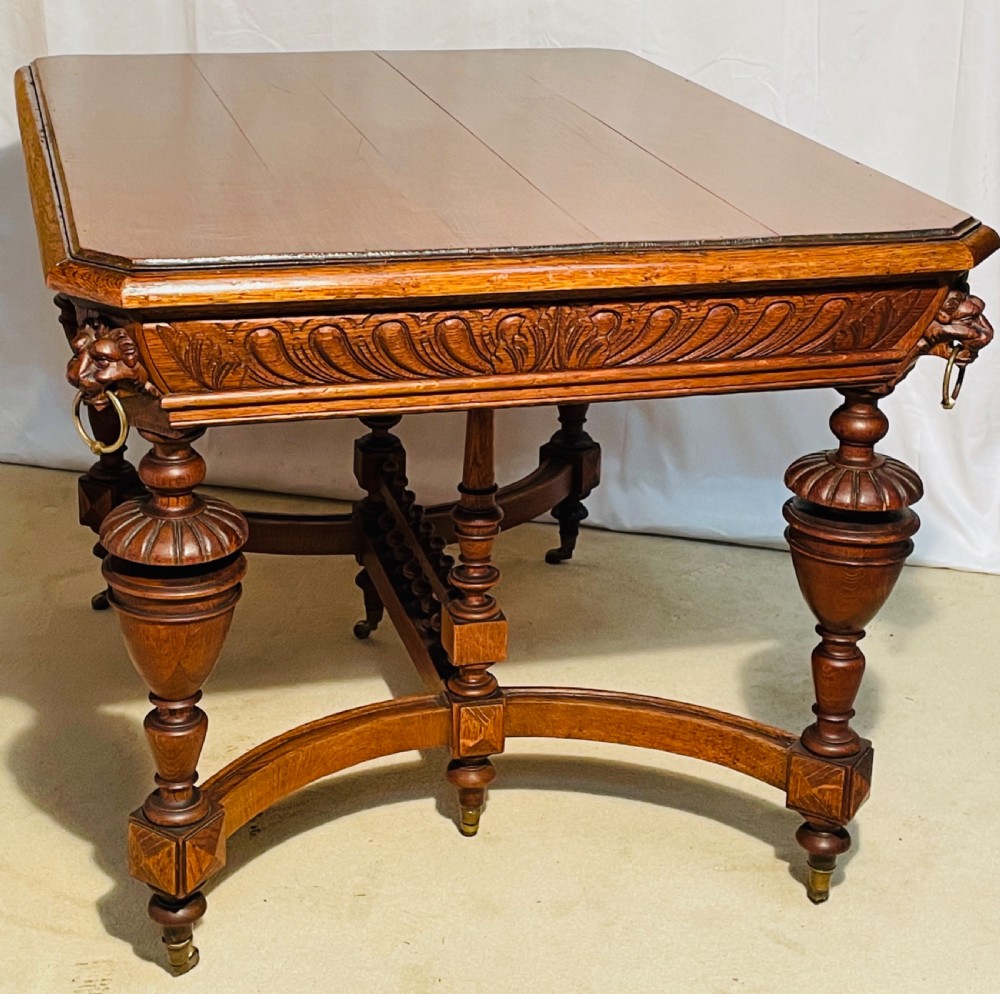 A Victorian Gothic honey oak centre table featuring carved aprons, corner lion mask motifs with brass rings, turned legs, and an arched cross-stretcher base on castors. View VII