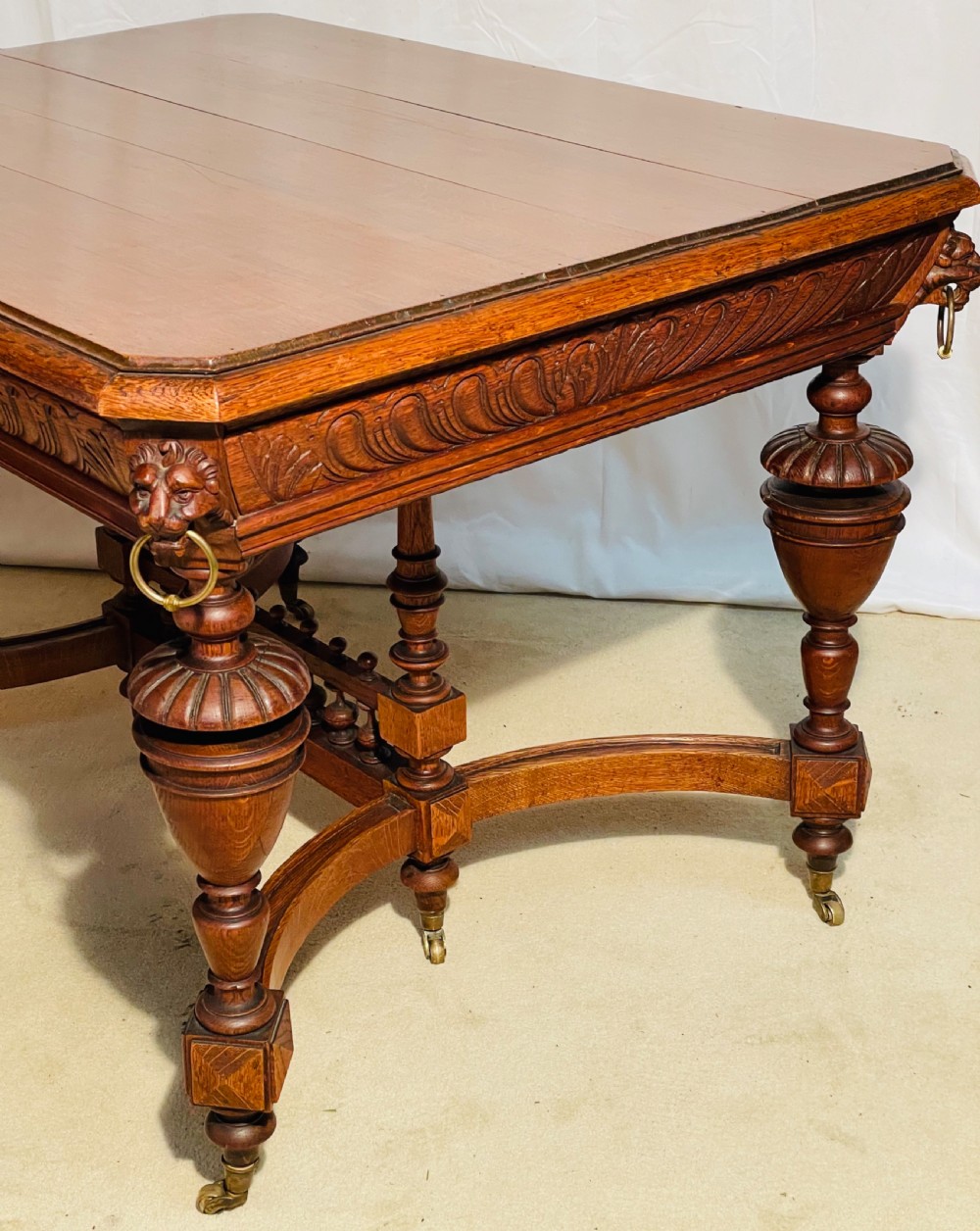 A Victorian Gothic honey oak centre table featuring carved aprons, corner lion mask motifs with brass rings, turned legs, and an arched cross-stretcher base on castors. View VIII