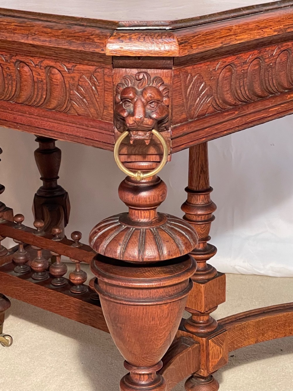 A Victorian Gothic honey oak centre table featuring carved aprons, corner lion mask motifs with brass rings, turned legs, and an arched cross-stretcher base on castors. View IX
