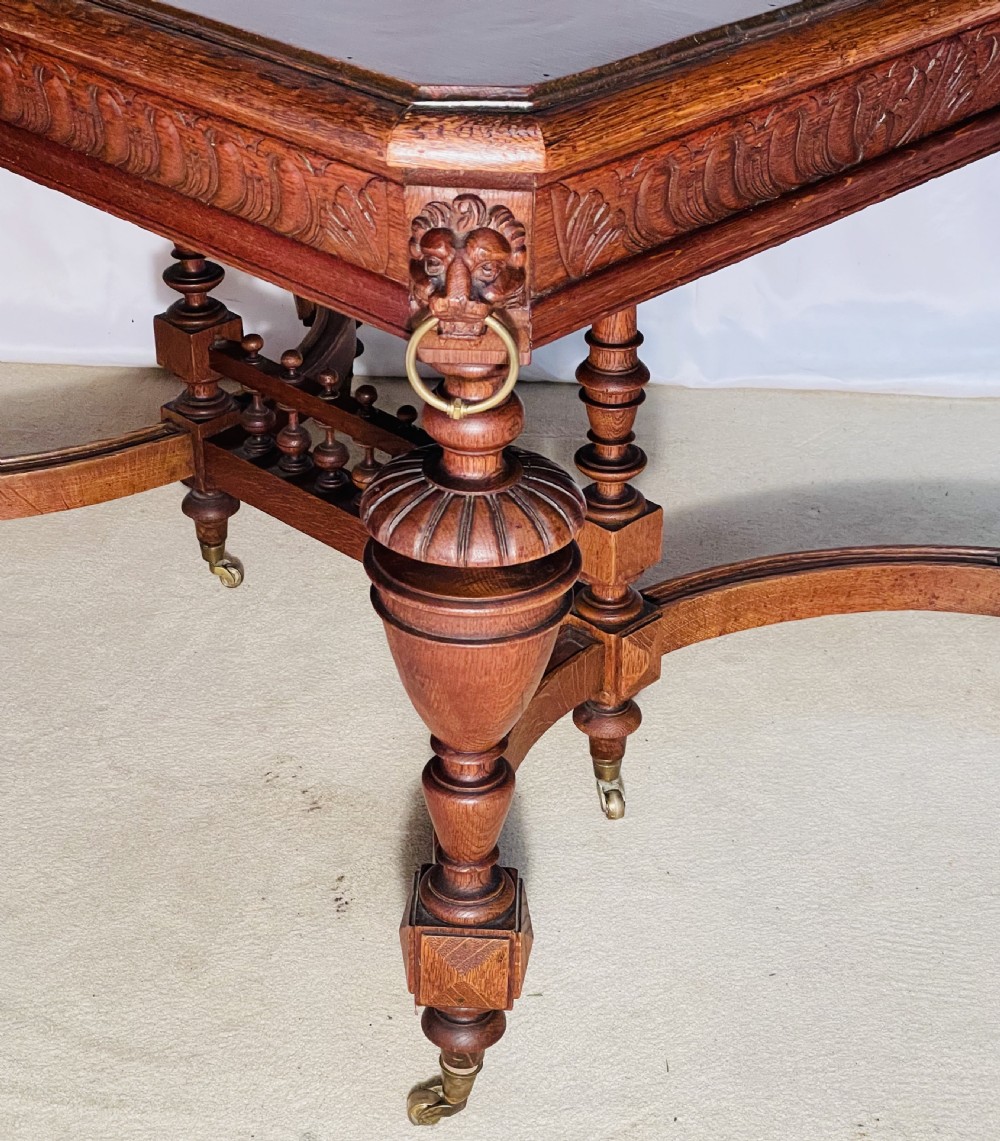 A Victorian Gothic honey oak centre table featuring carved aprons, corner lion mask motifs with brass rings, turned legs, and an arched cross-stretcher base on castors. View X