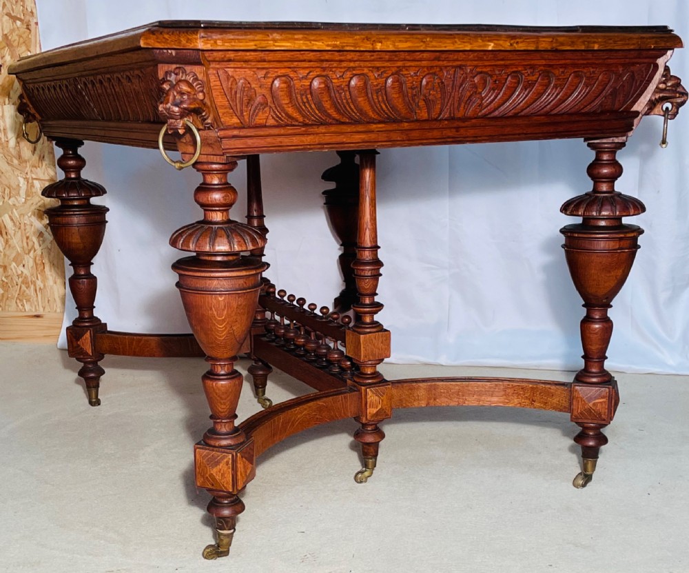 A Victorian Gothic honey oak centre table featuring carved aprons, corner lion mask motifs with brass rings, turned legs, and an arched cross-stretcher base on castors. View XI