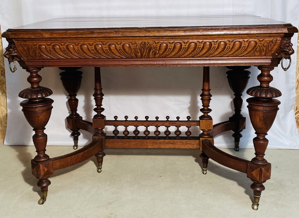 A Victorian Gothic honey oak centre table featuring carved aprons, corner lion mask motifs with brass rings, turned legs, and an arched cross-stretcher base on castors. View XII