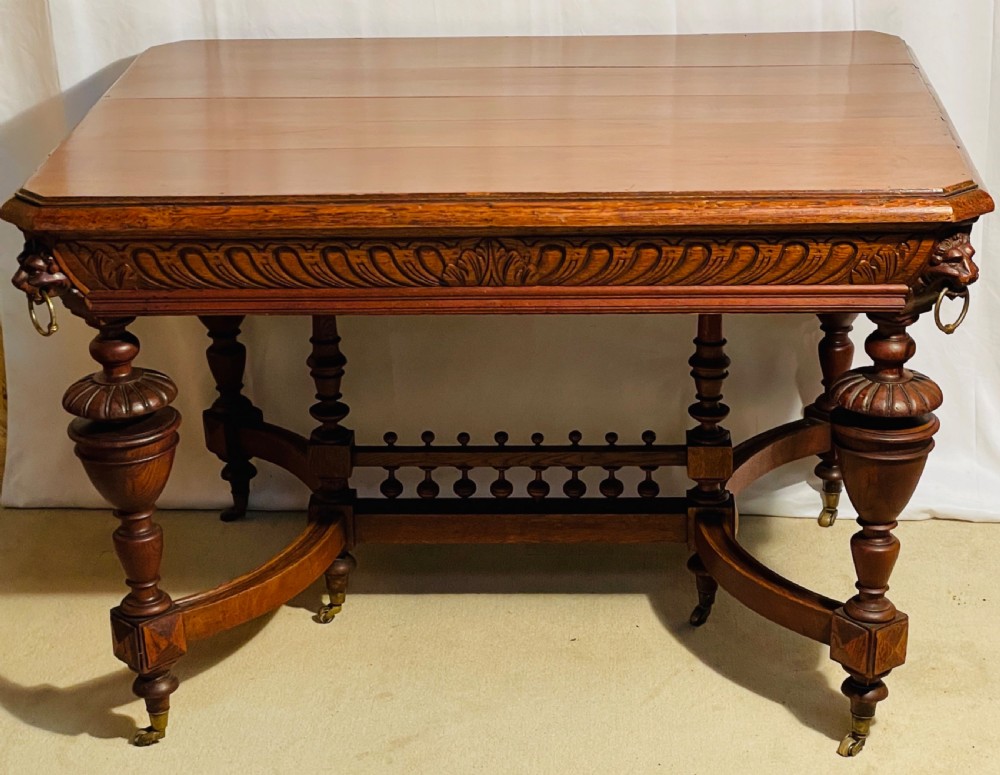 A Victorian Gothic honey oak centre table featuring carved aprons, corner lion mask motifs with brass rings, turned legs, and an arched cross-stretcher base on castors. View XIII
