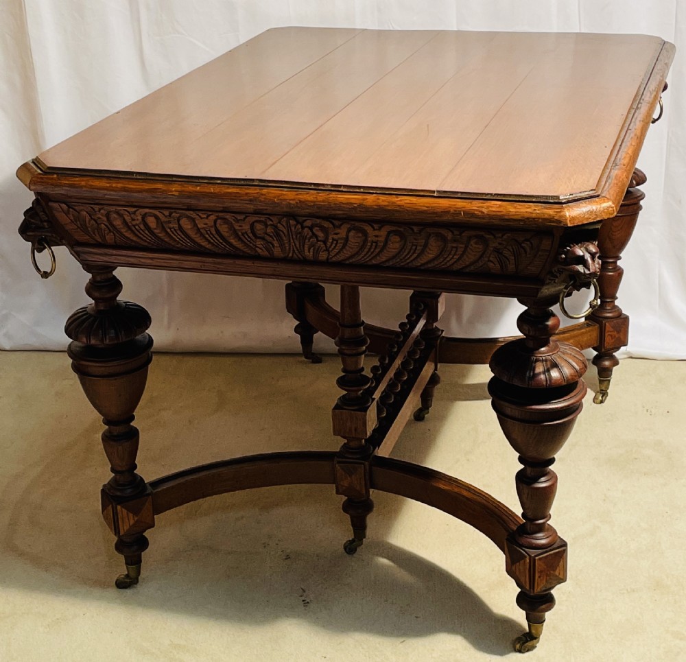 A Victorian Gothic honey oak centre table featuring carved aprons, corner lion mask motifs with brass rings, turned legs, and an arched cross-stretcher base on castors. View XIV