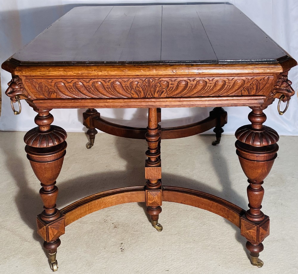 A Victorian Gothic honey oak centre table featuring carved aprons, corner lion mask motifs with brass rings, turned legs, and an arched cross-stretcher base on castors. View XV