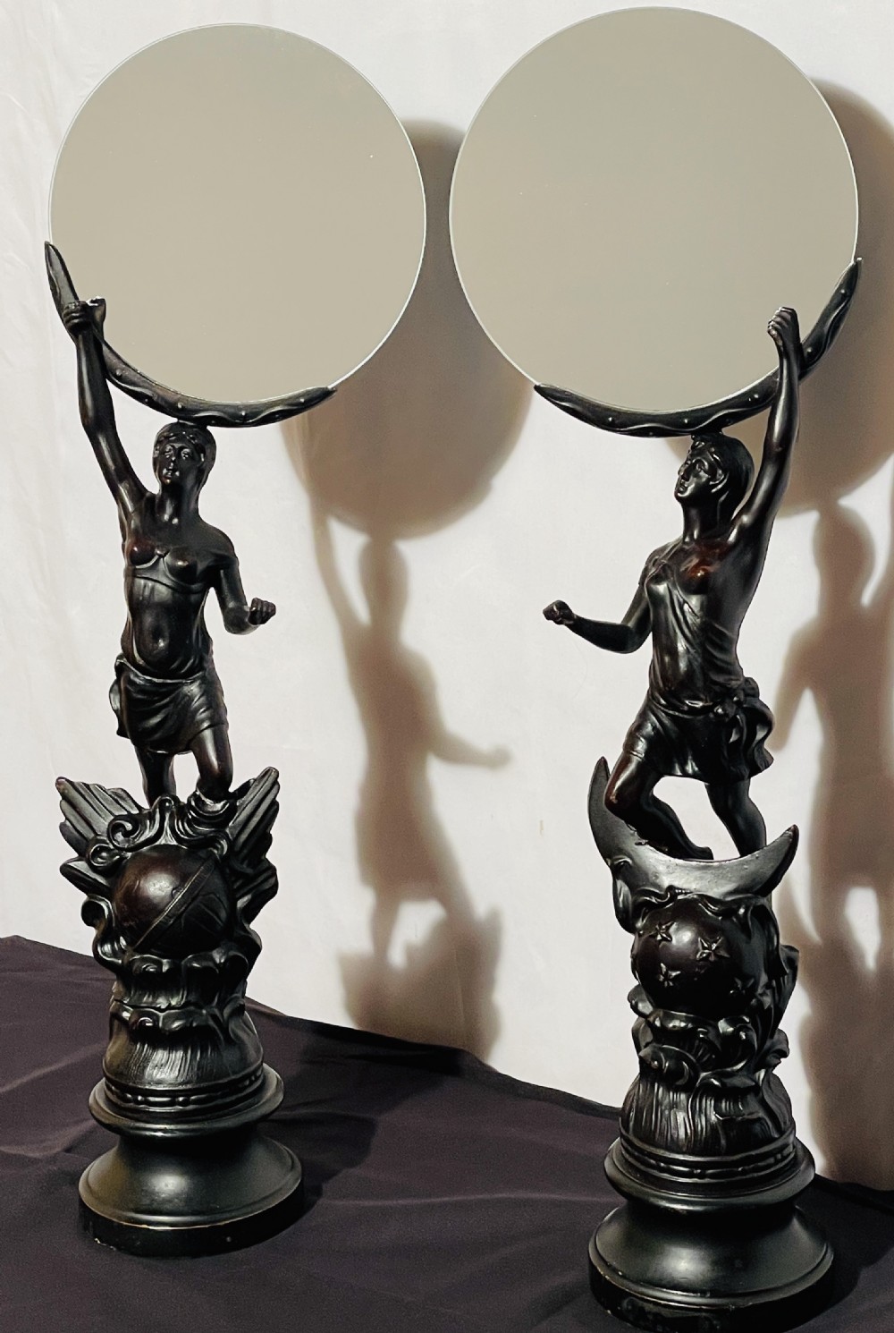 A pair of dark patinated spelter table mirrors featuring classical female figures standing on celestial-themed bases holding circular glass mirrors above their heads.