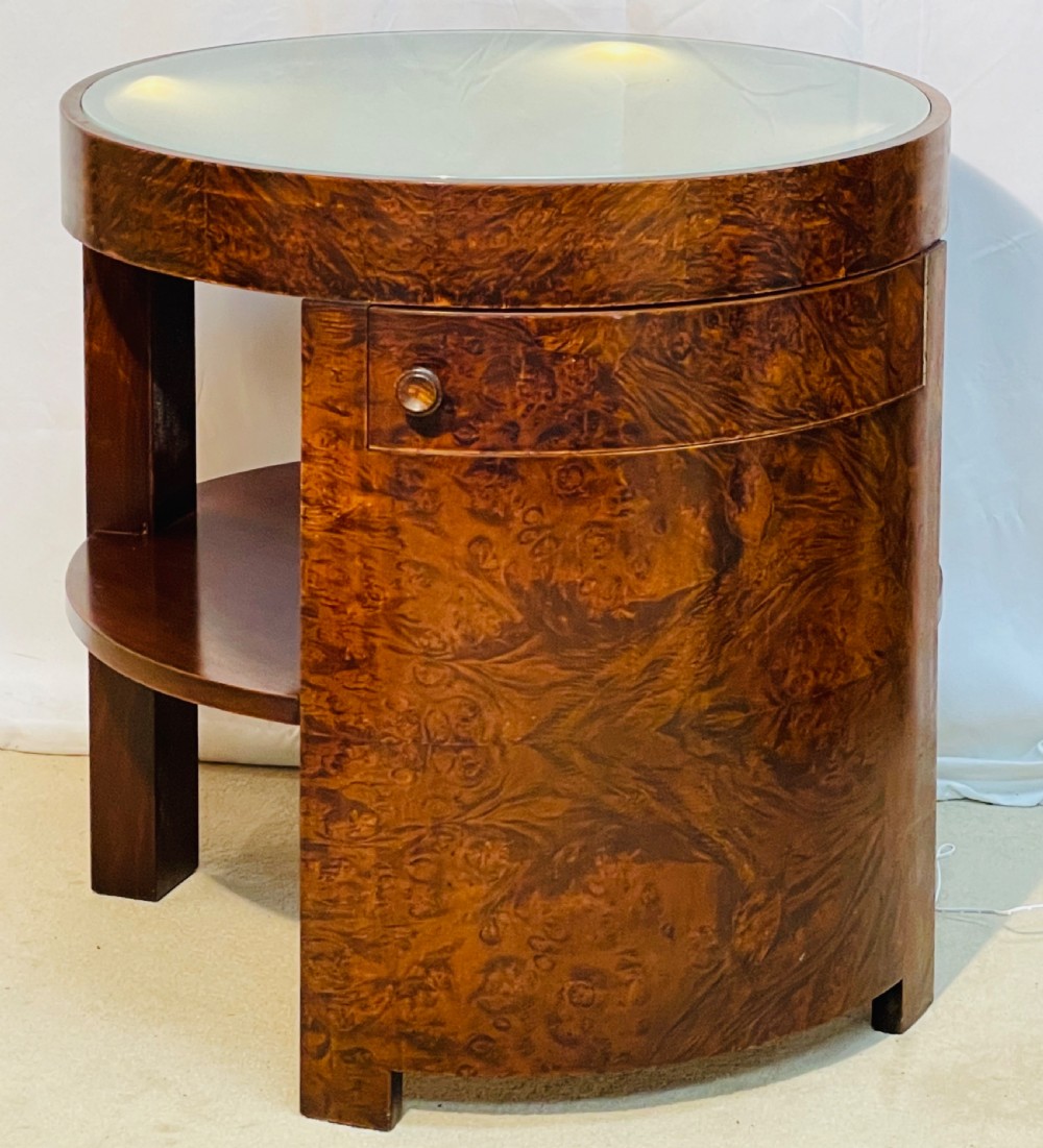 A circular Art Deco occasional table made from rich burr walnut veneer featuring an inset illuminated frosted glass top, curved side door, and lower shelf.