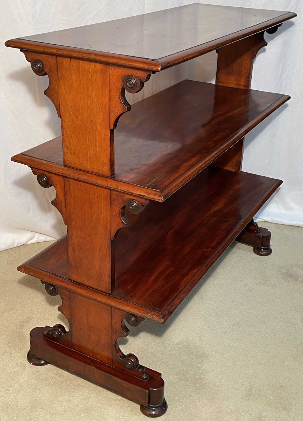 A three-tiered William IV mahogany dumb waiter featuring carved scroll supports with carved roundel details and a polished finish.