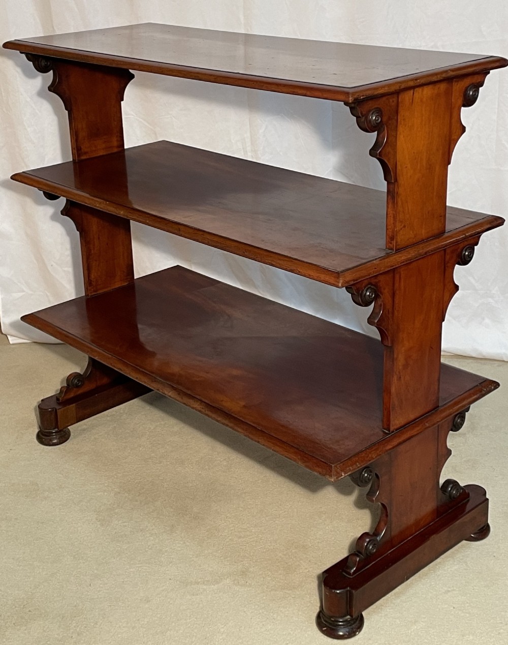 A three-tiered William IV mahogany dumb waiter featuring carved scroll supports with carved roundel details and a polished finish. View II