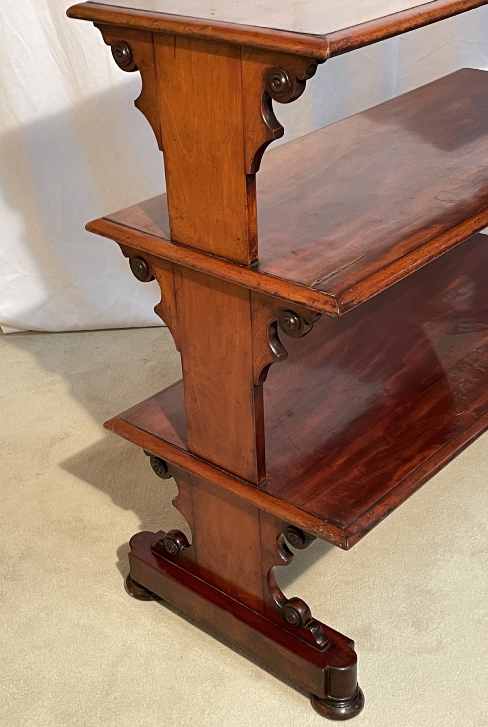 A three-tiered William IV mahogany dumb waiter featuring carved scroll supports with carved roundel details and a polished finish. View VII