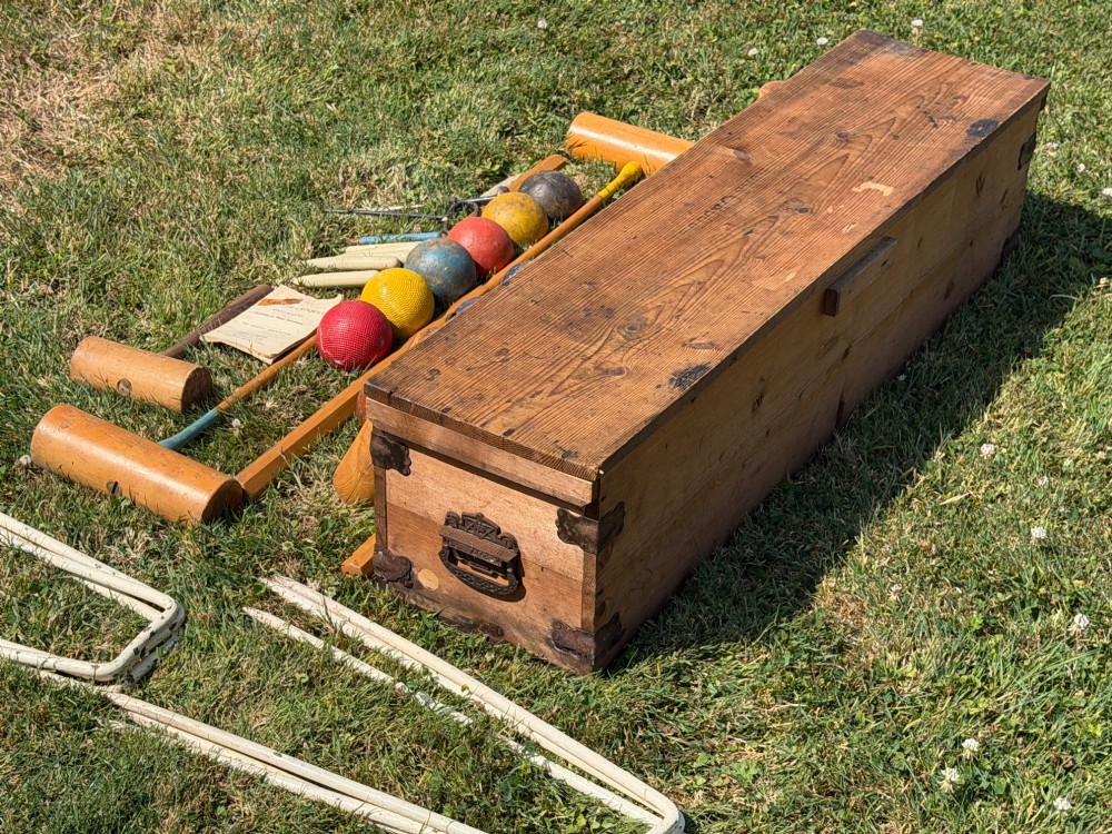 An open pine storage box with a Slazenger printed label displaying four wooden croquet mallets, coloured balls, metal hoops, and wooden pegs. View V