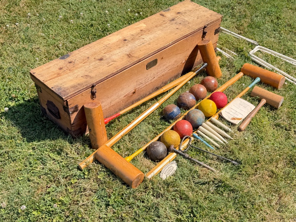 An open pine storage box with a Slazenger printed label displaying four wooden croquet mallets, coloured balls, metal hoops, and wooden pegs. View X