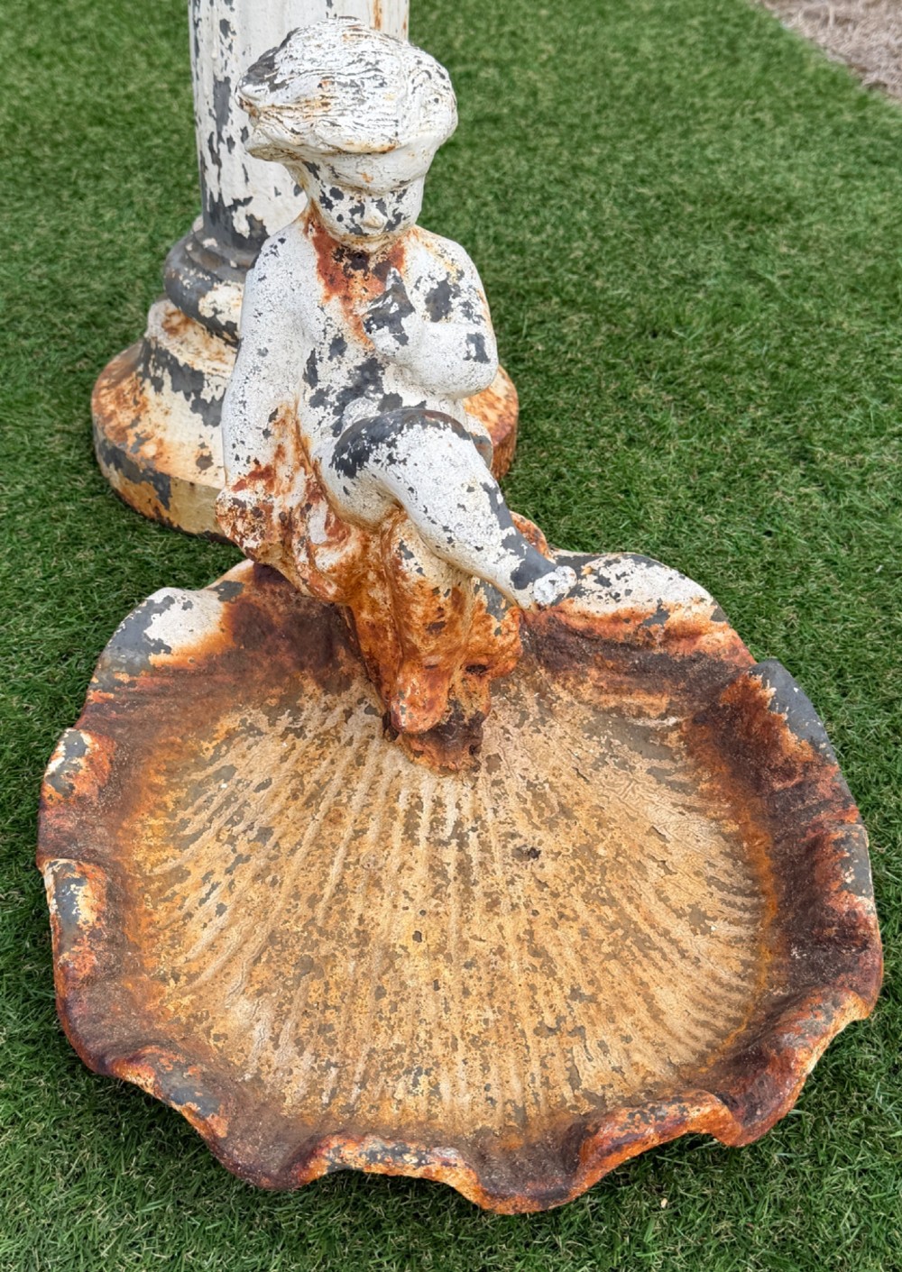 A late Victorian cast iron bird bath featuring a fluted column base and a scallop shell basin topped with a seated cherub figure, showing weathered paint and rust oxidisation. View II