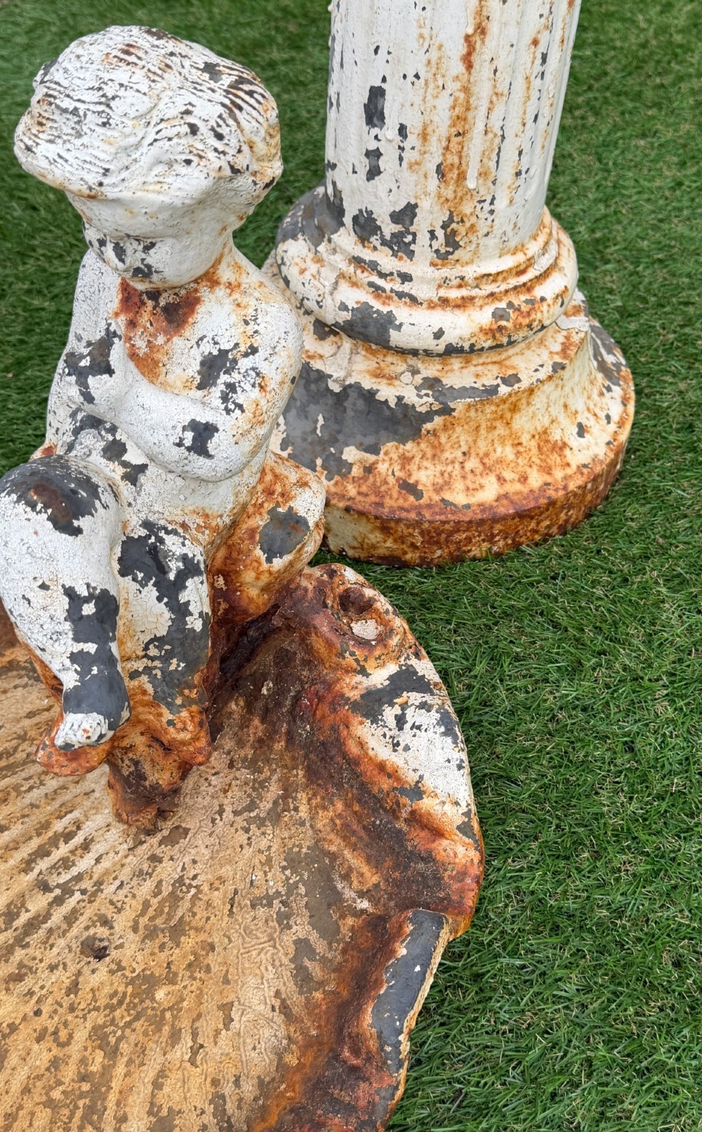 A late Victorian cast iron bird bath featuring a fluted column base and a scallop shell basin topped with a seated cherub figure, showing weathered paint and rust oxidisation. View III