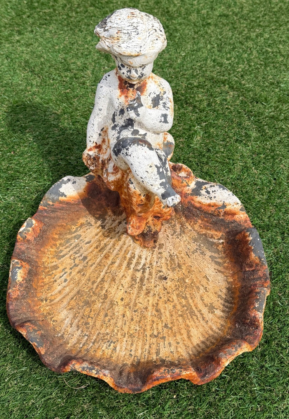 A late Victorian cast iron bird bath featuring a fluted column base and a scallop shell basin topped with a seated cherub figure, showing weathered paint and rust oxidisation. View V