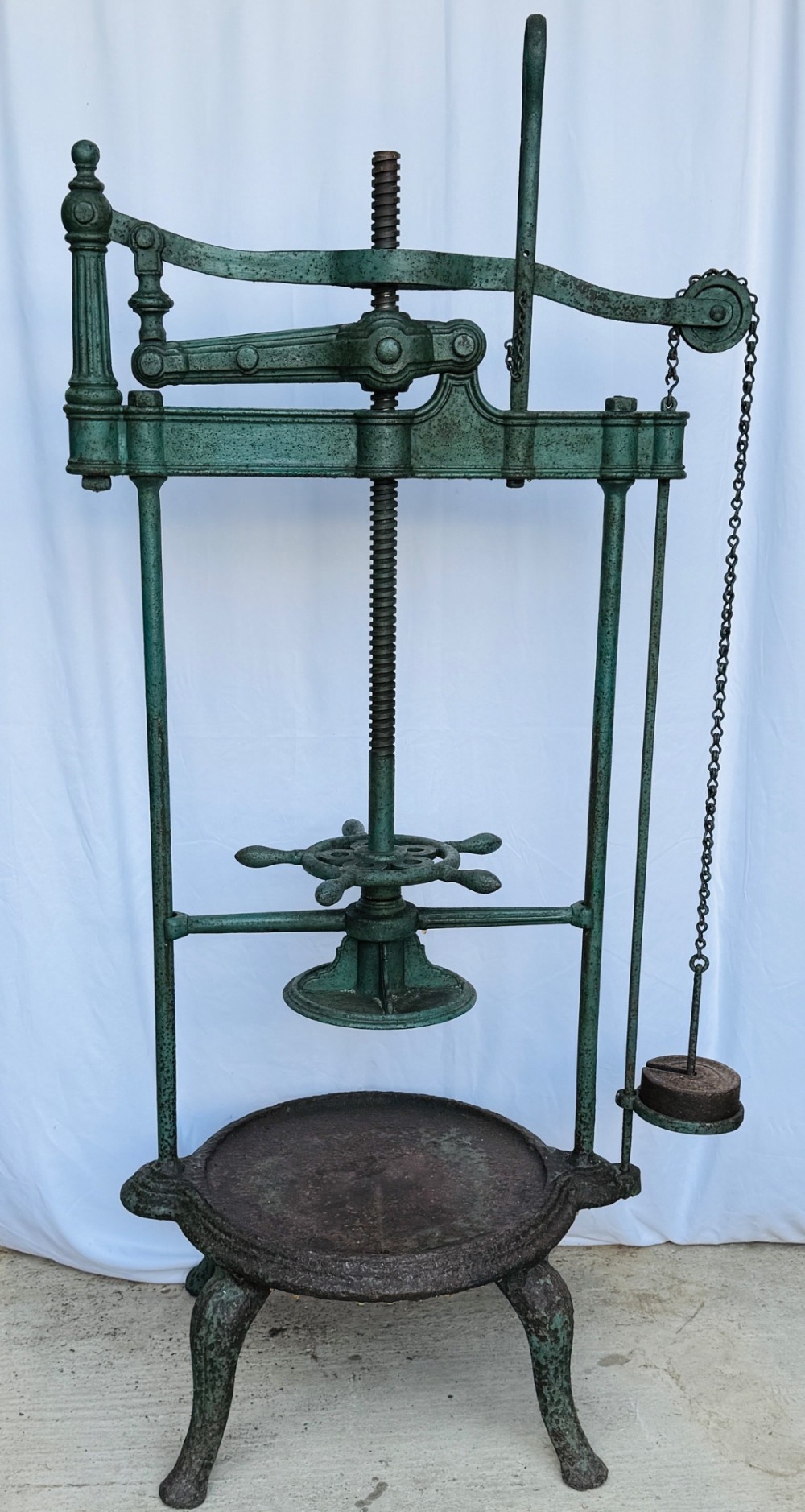 A Victorian patinated green cast iron and forged steel cheese press featuring a threaded screw mechanism, wheel handle, lever arm, chain, counterweight, and tripod legs. View VI