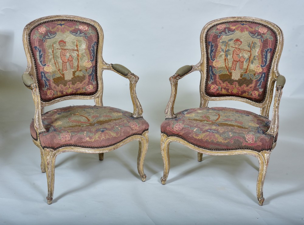 A pair of late 18th-century French painted wood fauteuil armchairs featuring curved cabriole legs and hand-stitched floral and figurative needlework upholstery.
