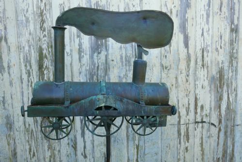 fig 1. A nineteenth-century copper weather vane shaped like a steam locomotive train with verdigris patina, featuring directional cardinal points on a cast iron stand.